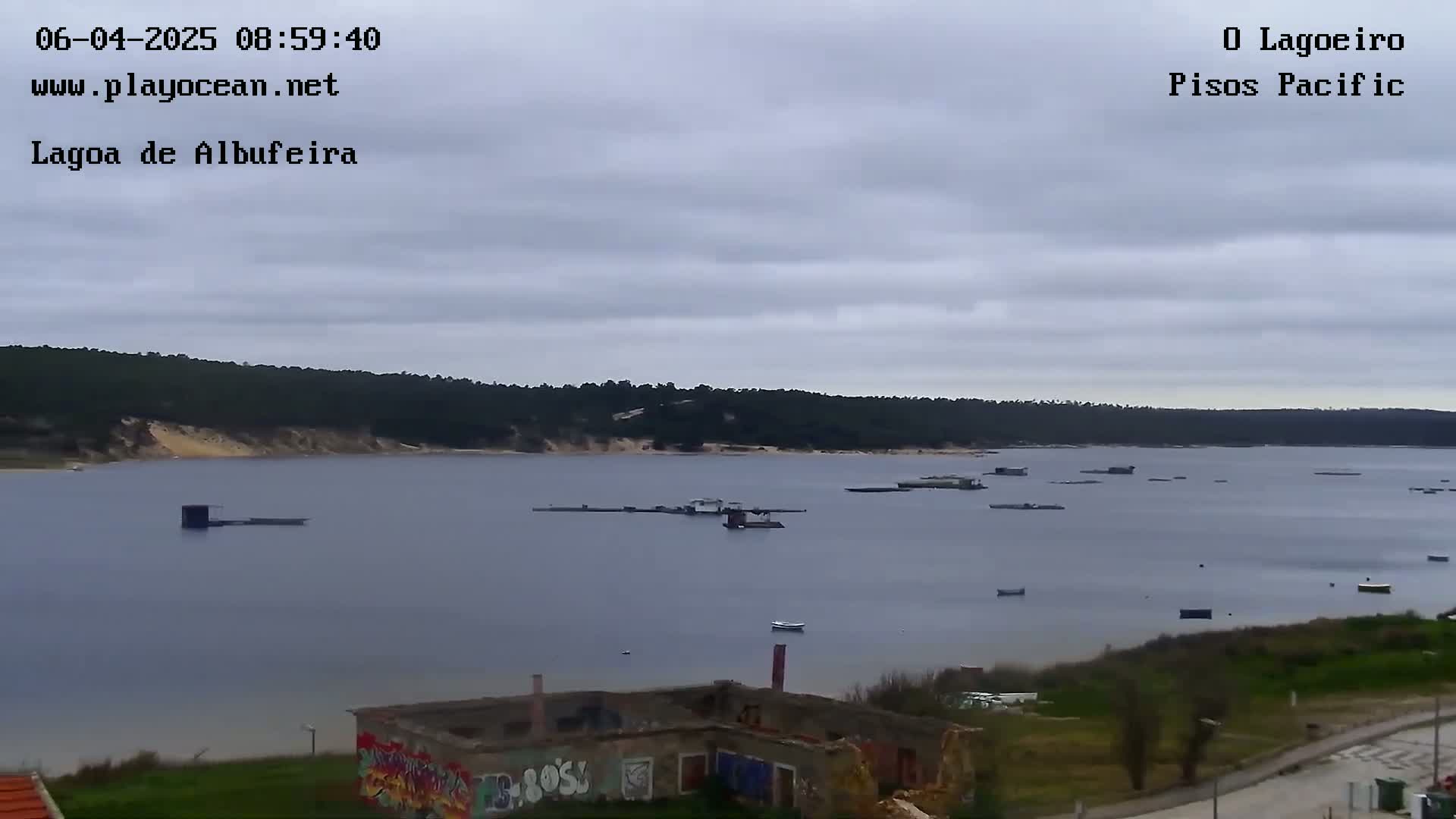 A calm body of water with numerous small structures and a few boats is seen under an overcast sky, with a wooded shoreline in the background.