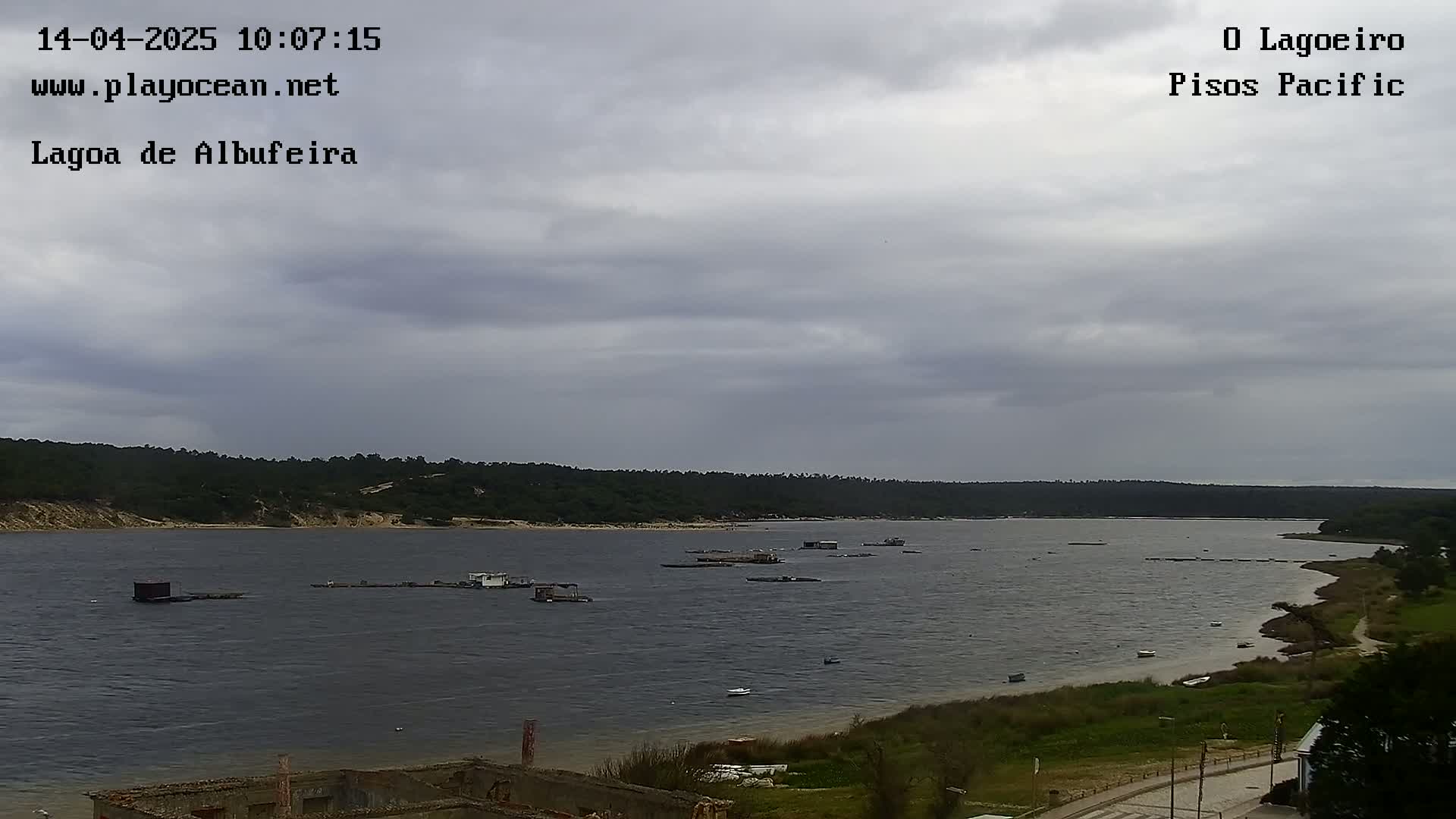 A body of water with several small structures and boats is visible under a cloudy sky, with a line of trees and vegetation along the far shore.
