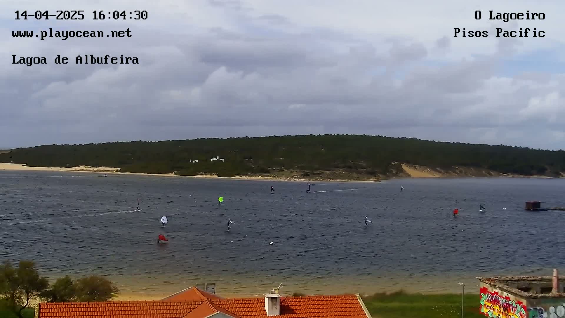 Several windsurfers are on a body of water under a cloudy sky, with a sandy shore and green hillside in the background.