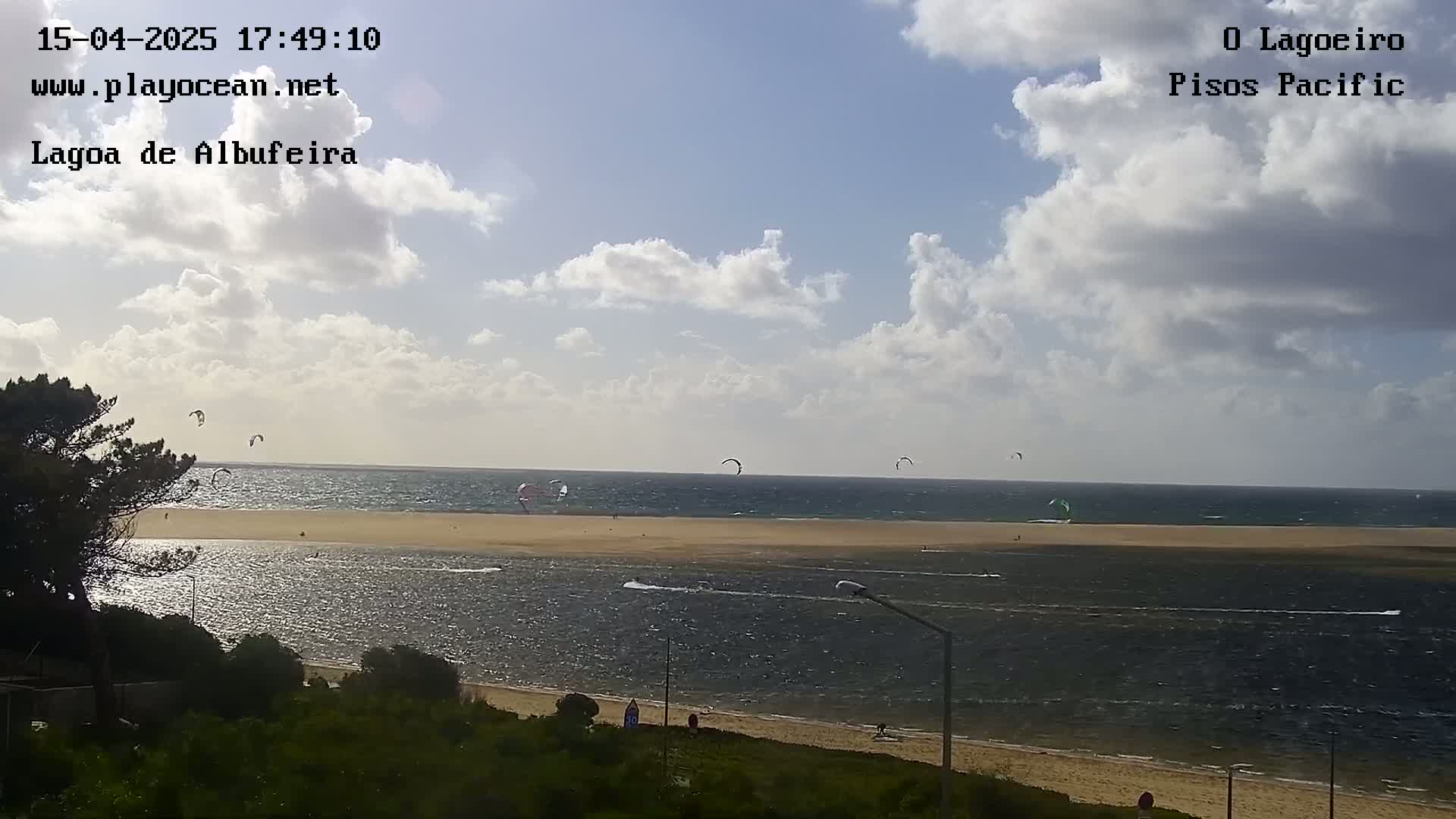 A partly cloudy day shows several kitesurfers enjoying a sandy beach and calm water.