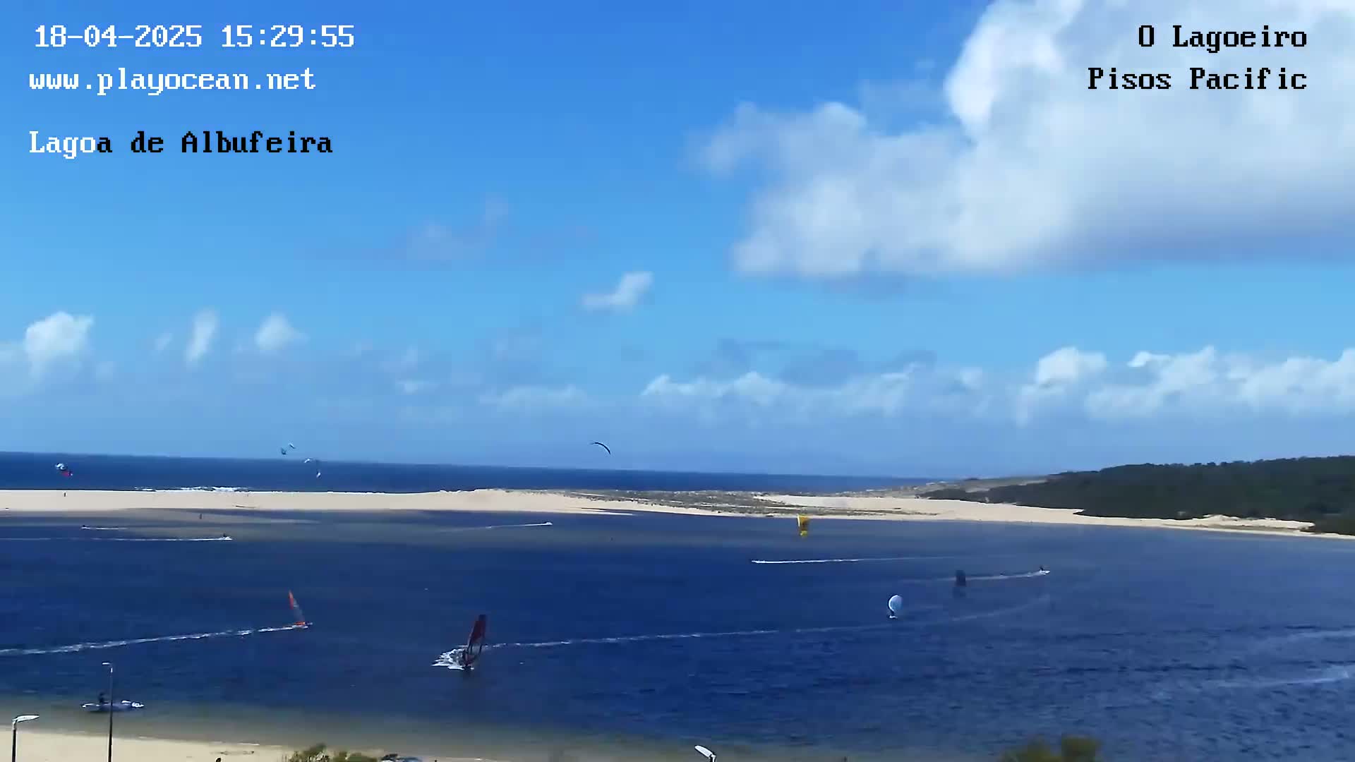 A mostly sunny day with a few clouds shows windsurfers and kitesurfers on a lagoon, with a sandy strip of land separating it from the ocean.