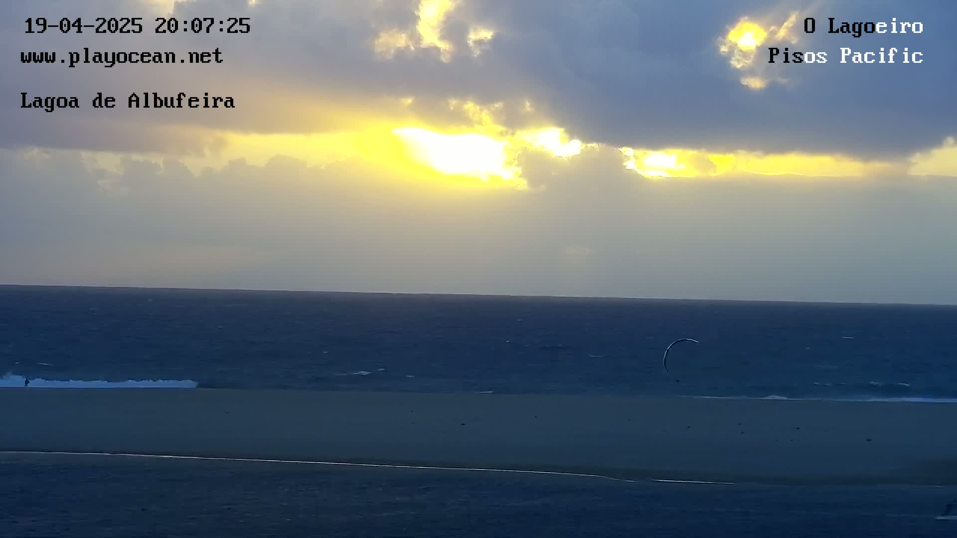 A partly cloudy sunset over a dark ocean and sandy beach, with a single kitesurfer visible in the distance.