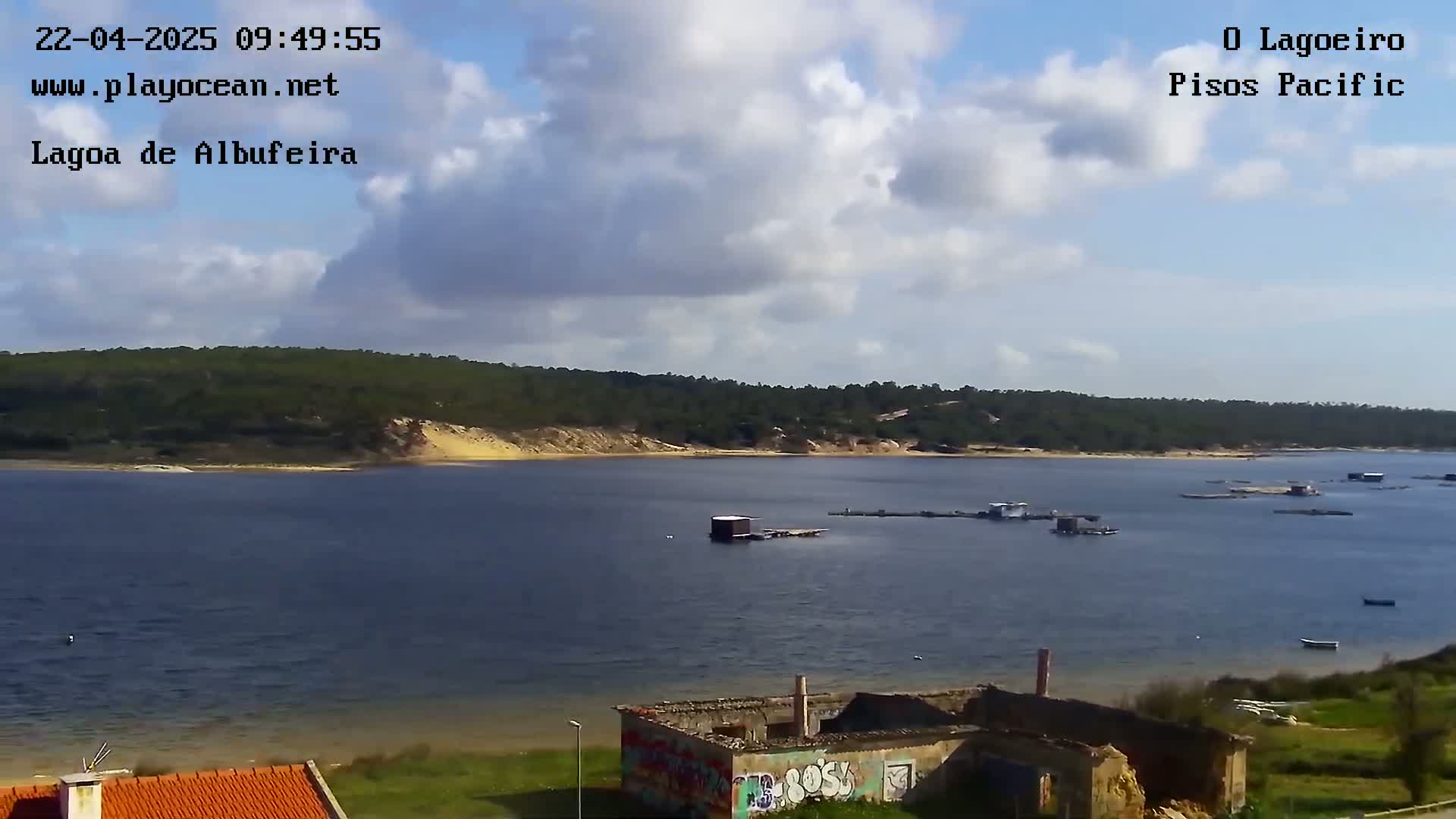 A partly cloudy day overlooks a calm body of water with several small structures on its surface, a ruined building in the foreground, and a forested shoreline in the background.