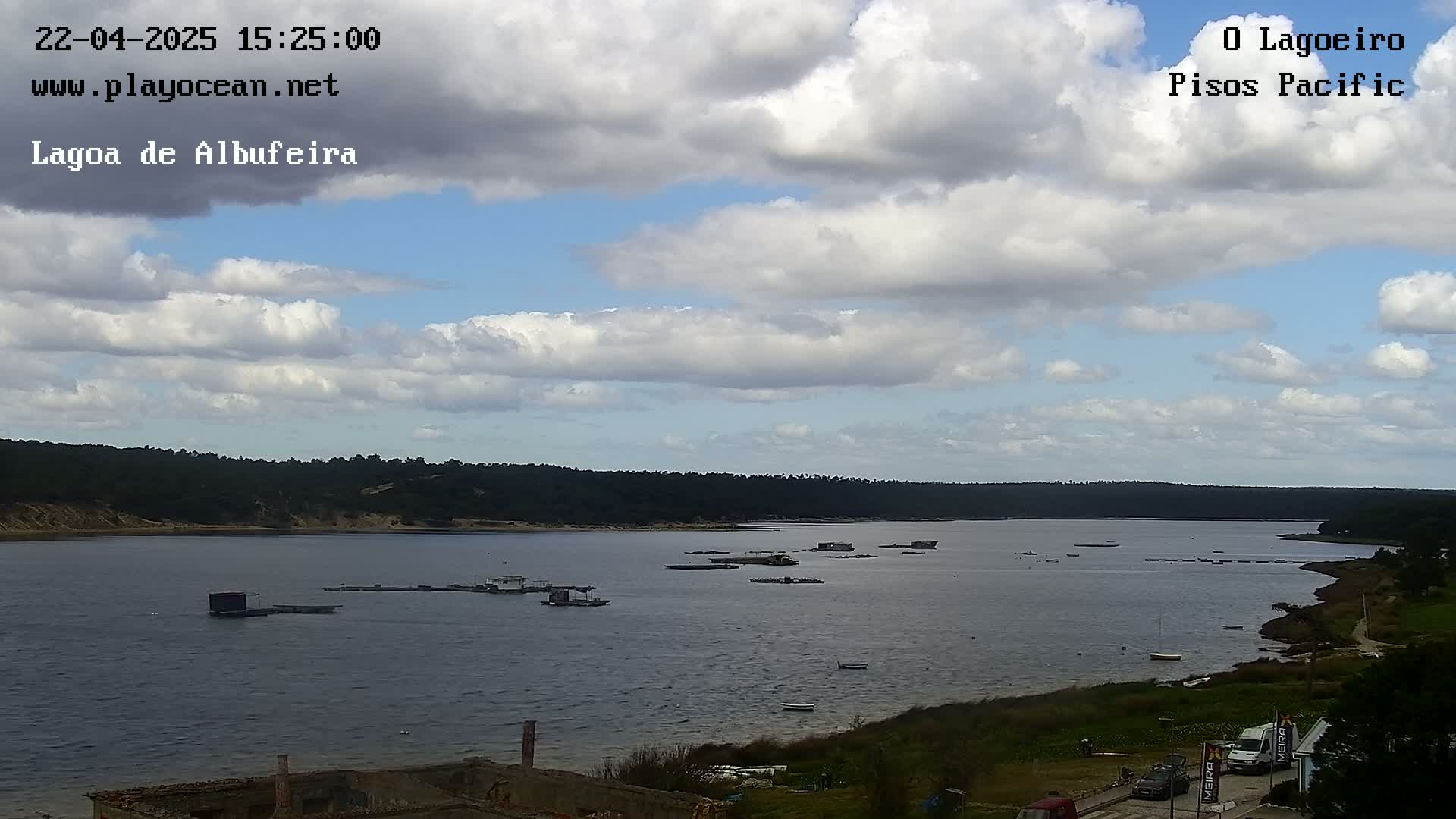 A partly cloudy day reveals a calm body of water with several small structures and boats scattered across its surface, backed by a line of dark green trees and sandy shore.