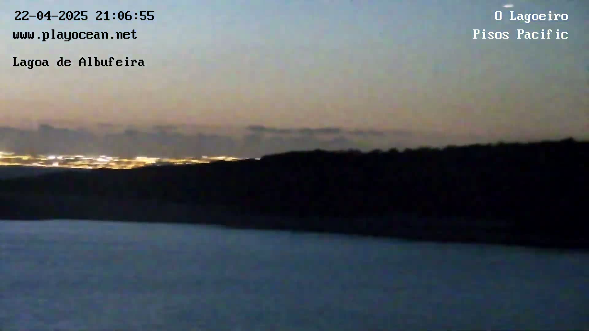 A dark, still body of water lies in the foreground, with a dark landmass and distant city lights visible under a twilight sky.