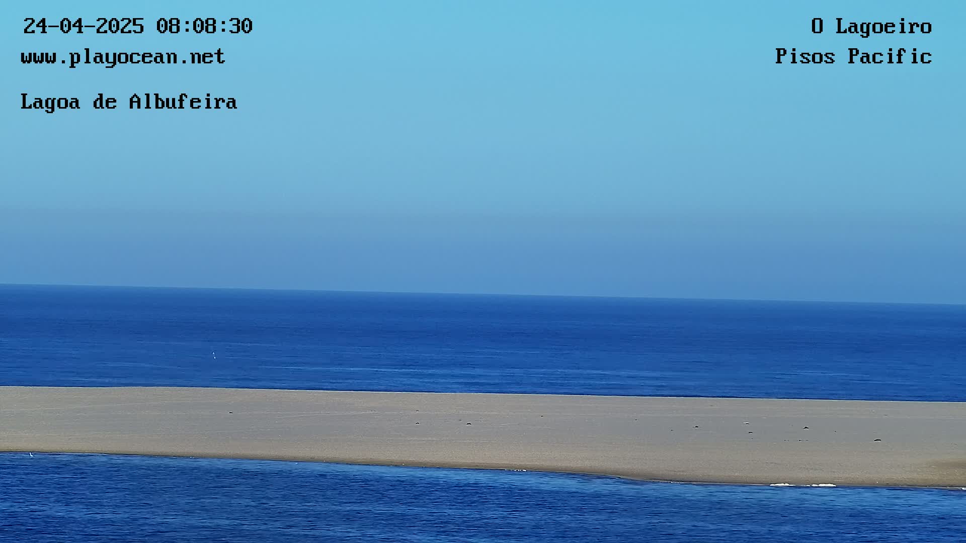 A long, sandy beach separates a calm, blue ocean from a similarly colored body of water under a clear blue sky.