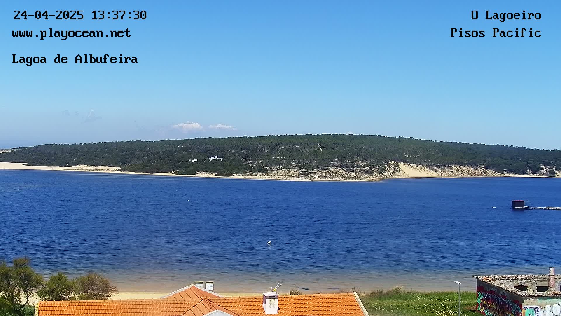 A calm, blue body of water stretches out before a sandy shoreline backed by a verdant hill under a mostly clear sky.
