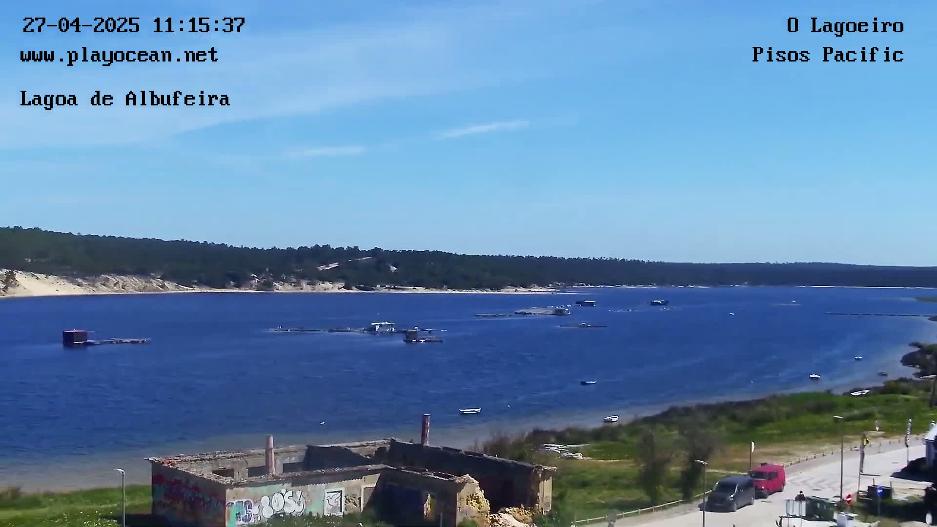 Under a clear blue sky, a calm blue lagoon stretches out, showing several small boats and structures on the water, with a ruined building and vehicles visible on the shore.