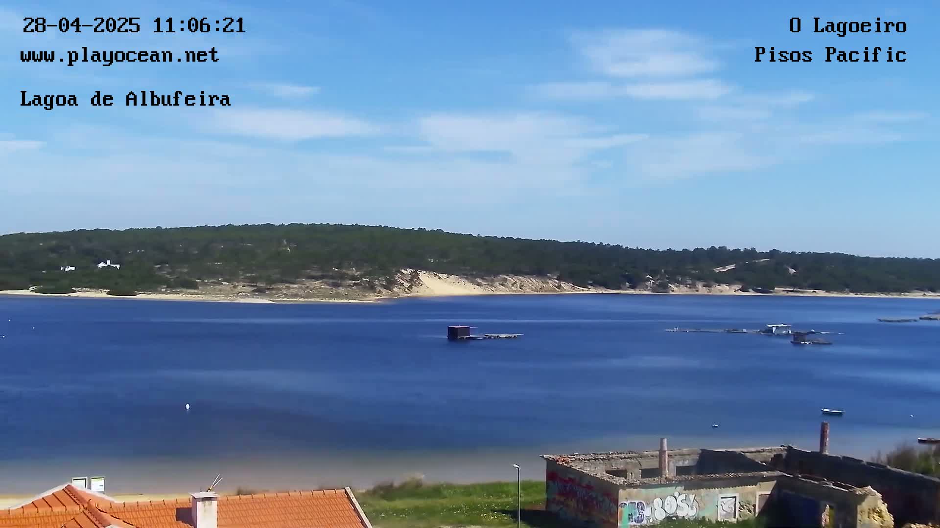 A calm, blue lagoon is seen under a mostly sunny sky, with a sandy shoreline and forested hills in the background, and some buildings and structures are visible in the foreground.