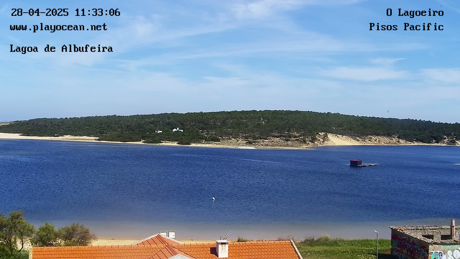 A body of blue water stretches to a sandy shoreline backed by green hills under a mostly clear, bright sky.
