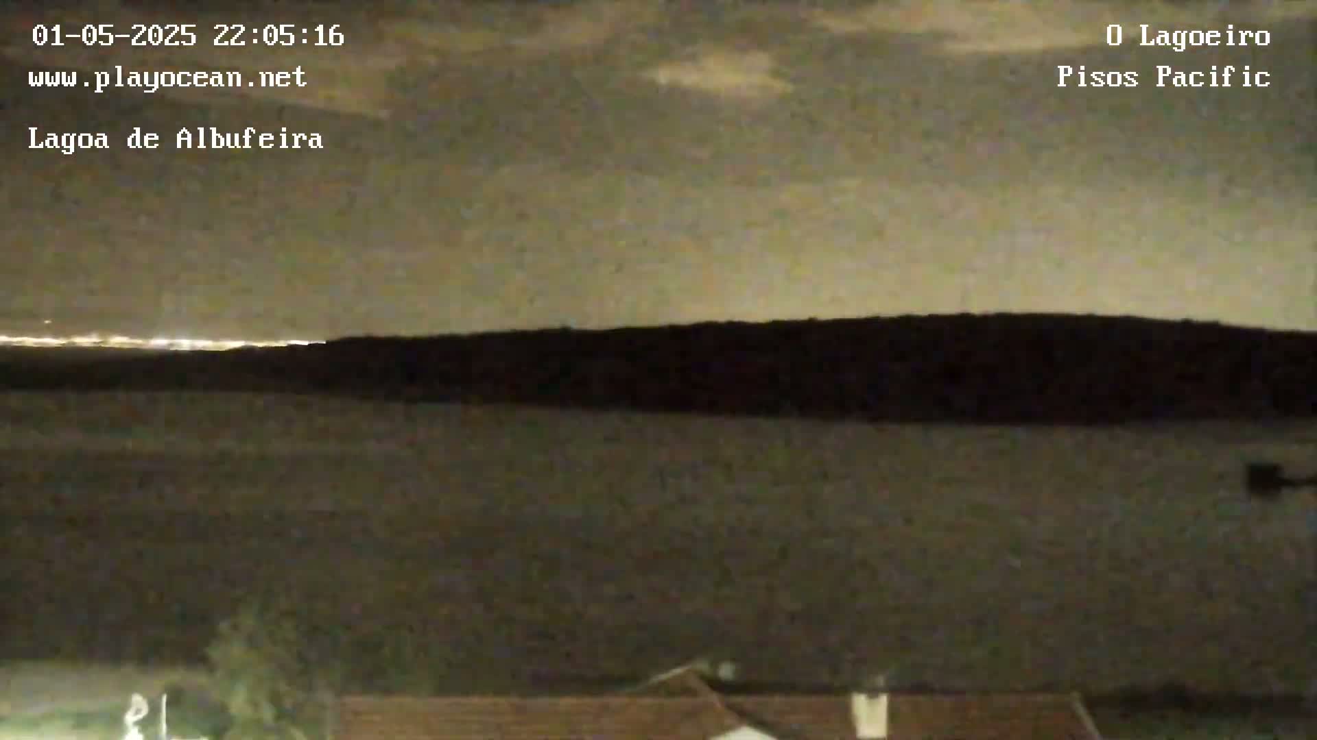 A nighttime view shows a dark body of water under a cloudy sky, with a distant line of lights on the horizon.