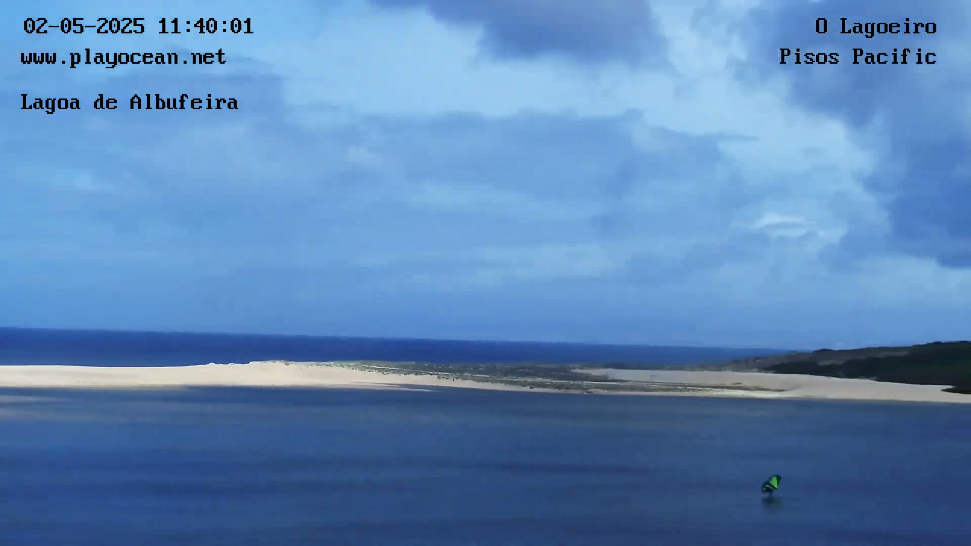 A windsurfer is visible on calm, blue water,  with a sandy shoreline and a partly cloudy sky.