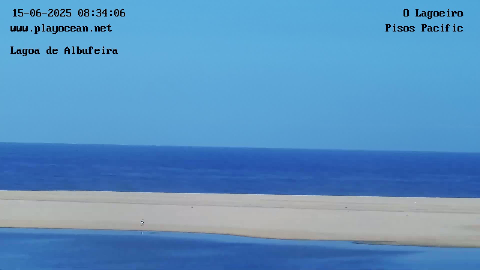 A long, wide expanse of sandy beach separates a calm, blue ocean from a tranquil body of water under a clear blue sky.