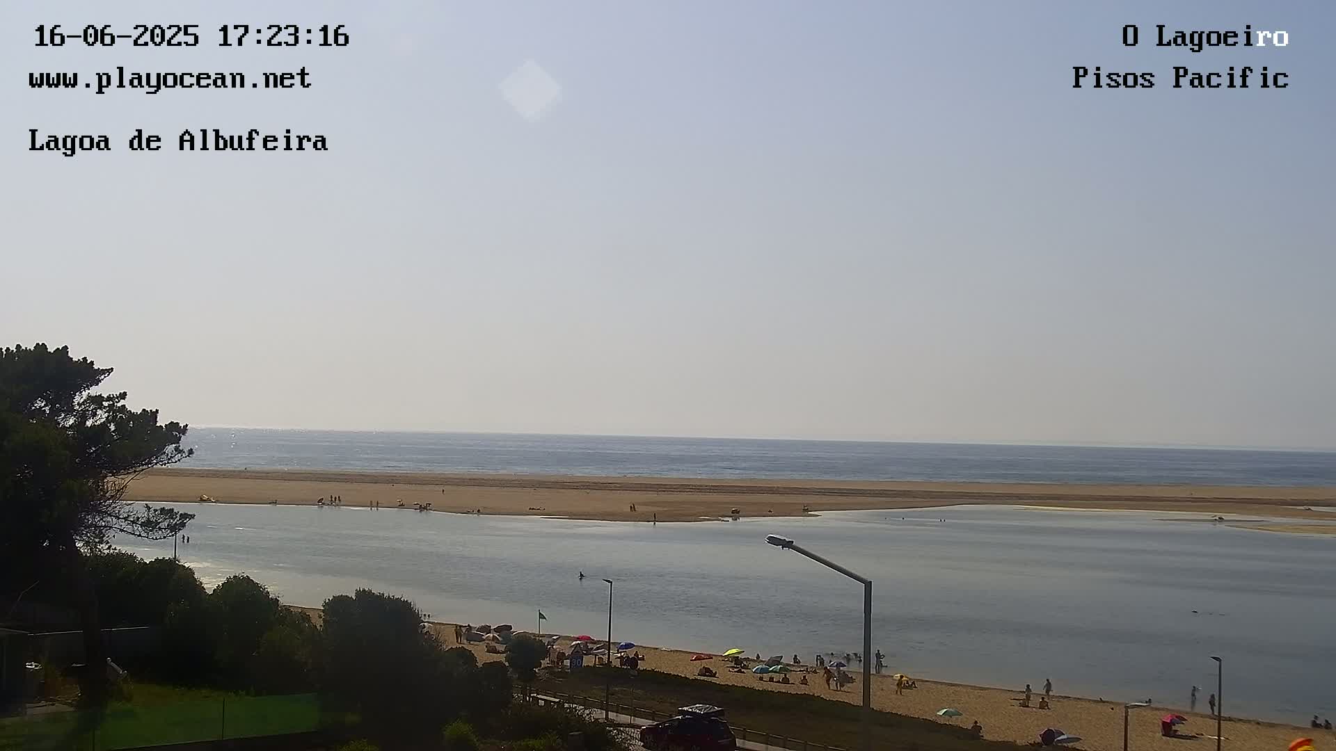 A calm ocean meets a sandy beach and lagoon where people are relaxing under umbrellas on a hazy day.