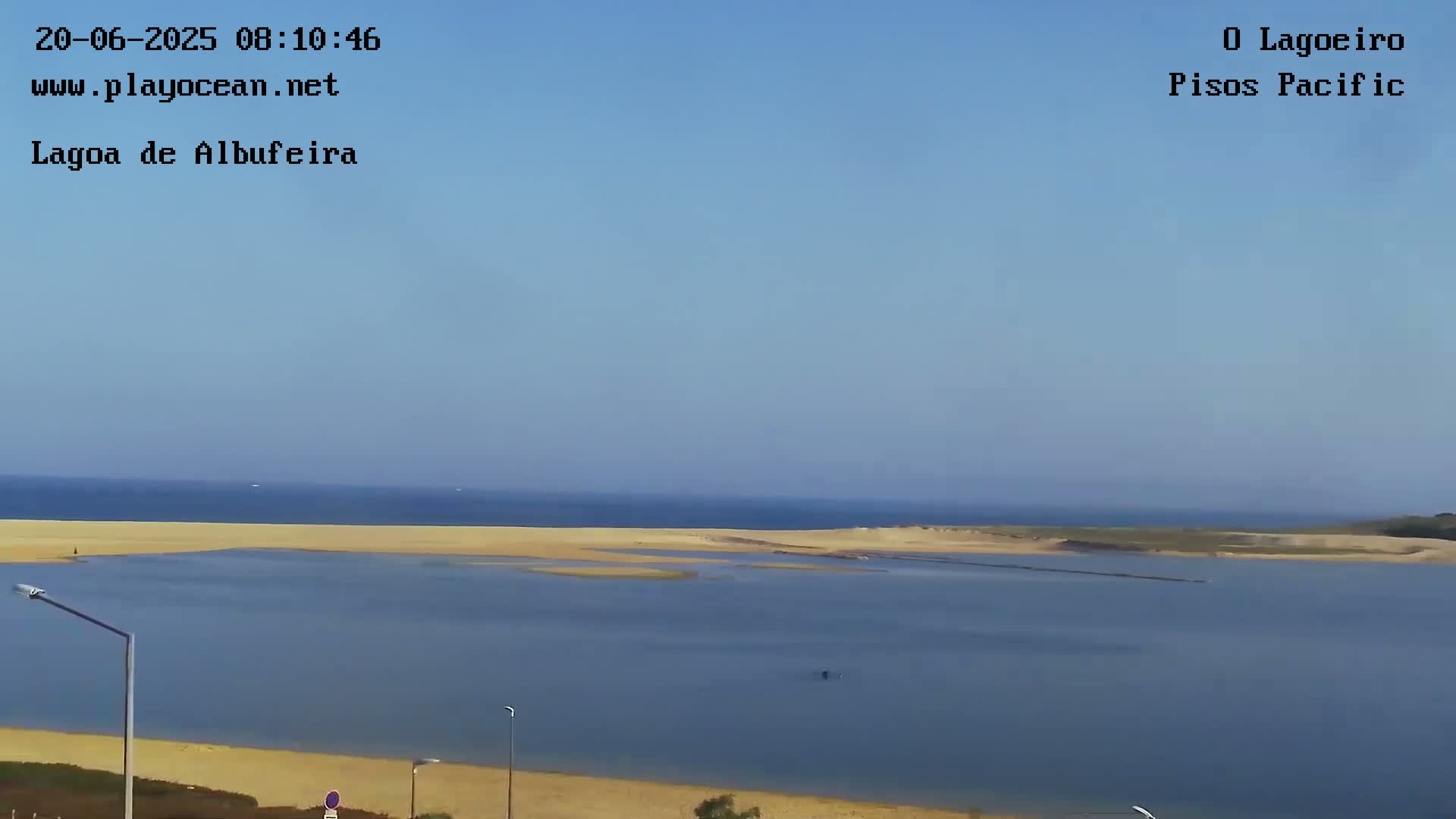 A calm, blue ocean stretches to the horizon under a clear sky, meeting a sandy beach that encloses a tranquil lagoon.