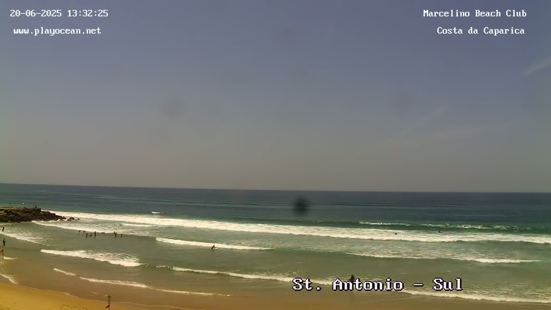 A mostly sunny day at a beach with gentle waves and a few people swimming and surfing.