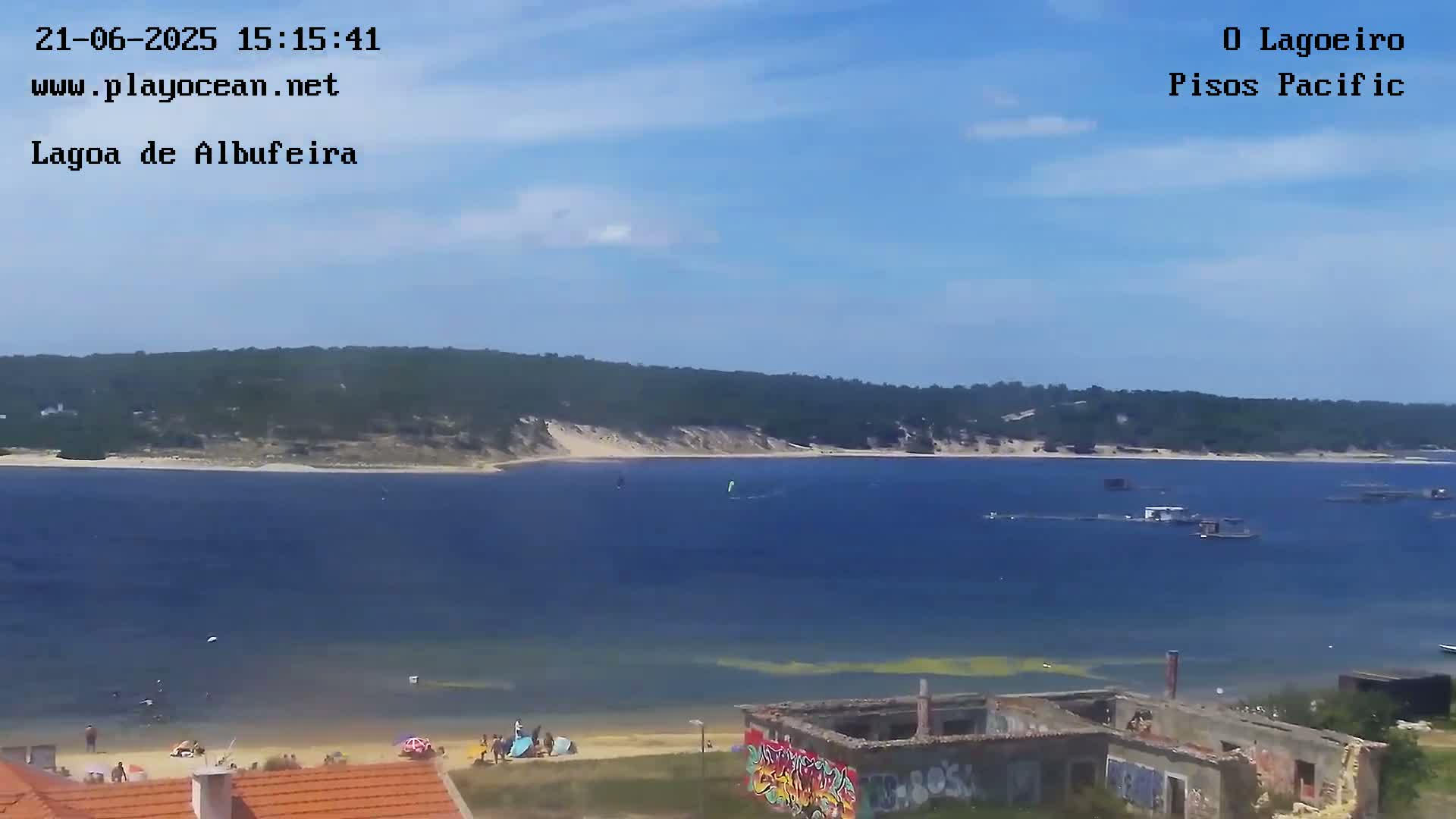 A sunny day shows a body of water with a sandy beach, a few people, and some structures, including a partially ruined building with graffiti.