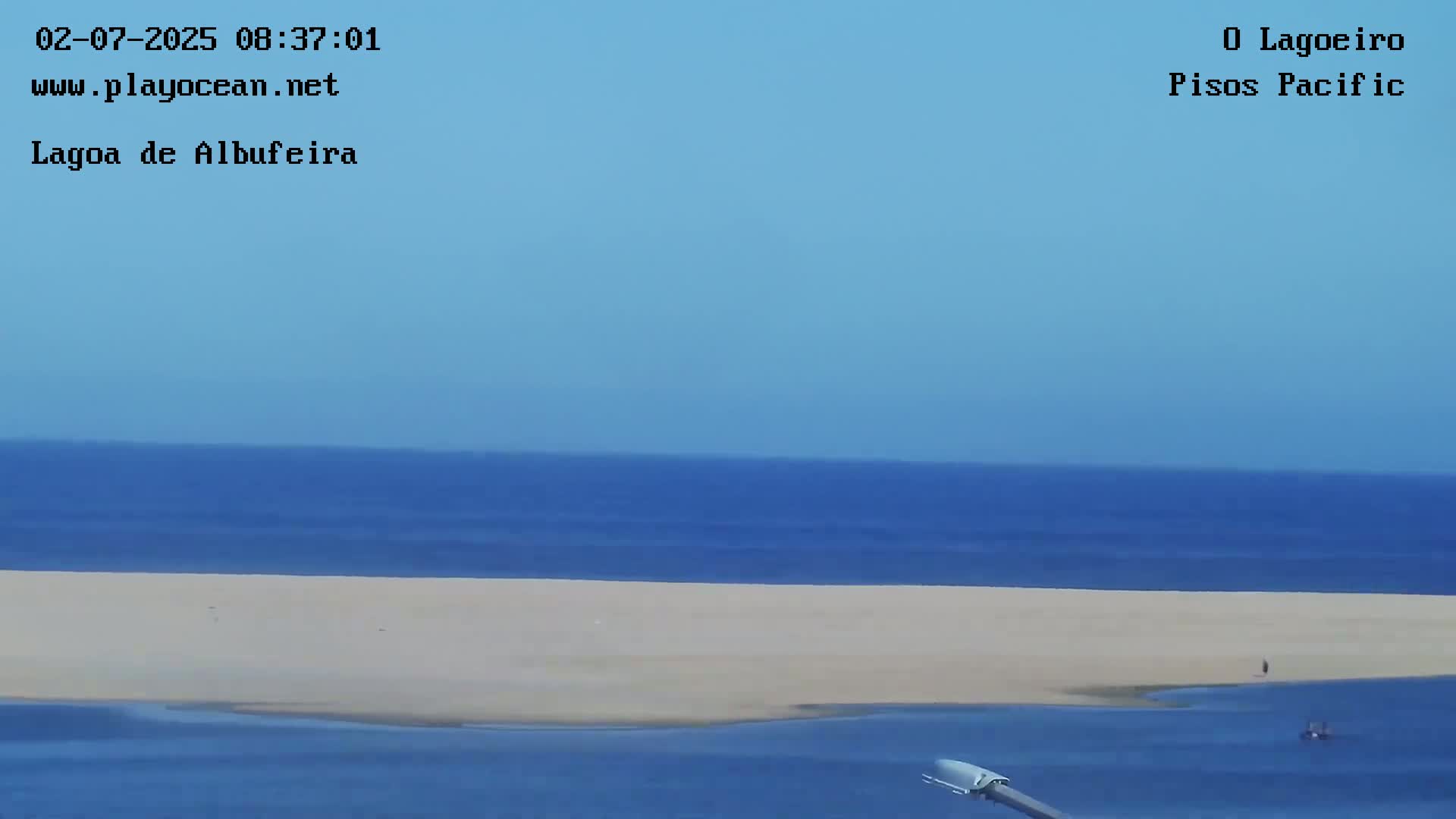 A long expanse of light-colored sand separates a calm, blue ocean from a darker blue body of water under a clear blue sky.