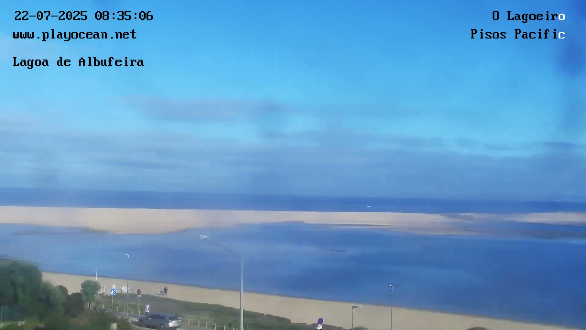 A mostly clear blue sky overlooks a calm body of water that meets a sandy beach, with a road and sparse vegetation visible in the foreground.