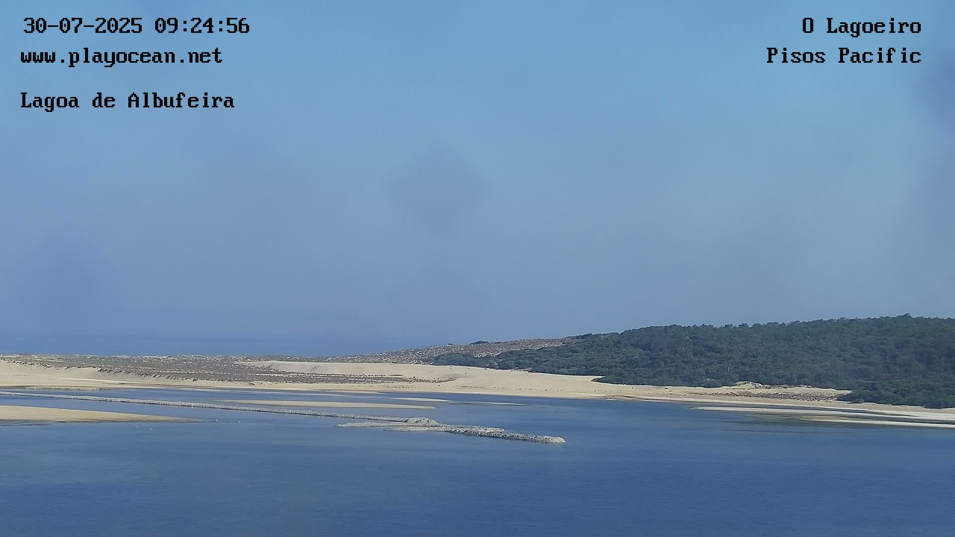 A tranquil lagoon is seen under a clear blue sky, with sandy shores and a line of dark green vegetation in the background.