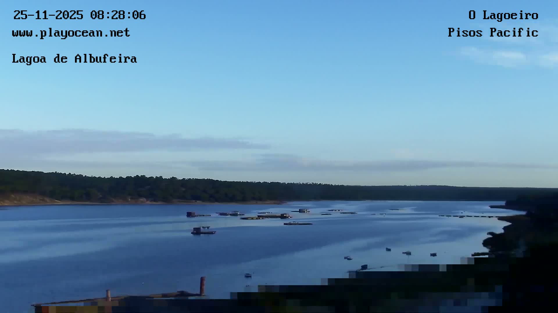 A tranquil blue body of water, possibly a lagoon, is shown dotted with numerous floating structures and boats, bordered by dark green forested shorelines under a clear blue sky with a few wispy clouds.