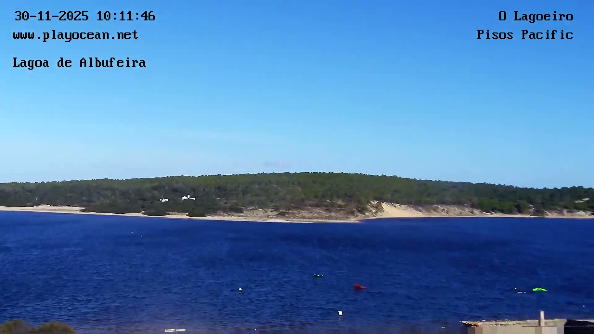 A serene clear blue sky hangs above a deep blue lagoon, bordered by a green, wooded shoreline with sandy stretches, and a few small colorful objects float on the water, all under sunny conditions.