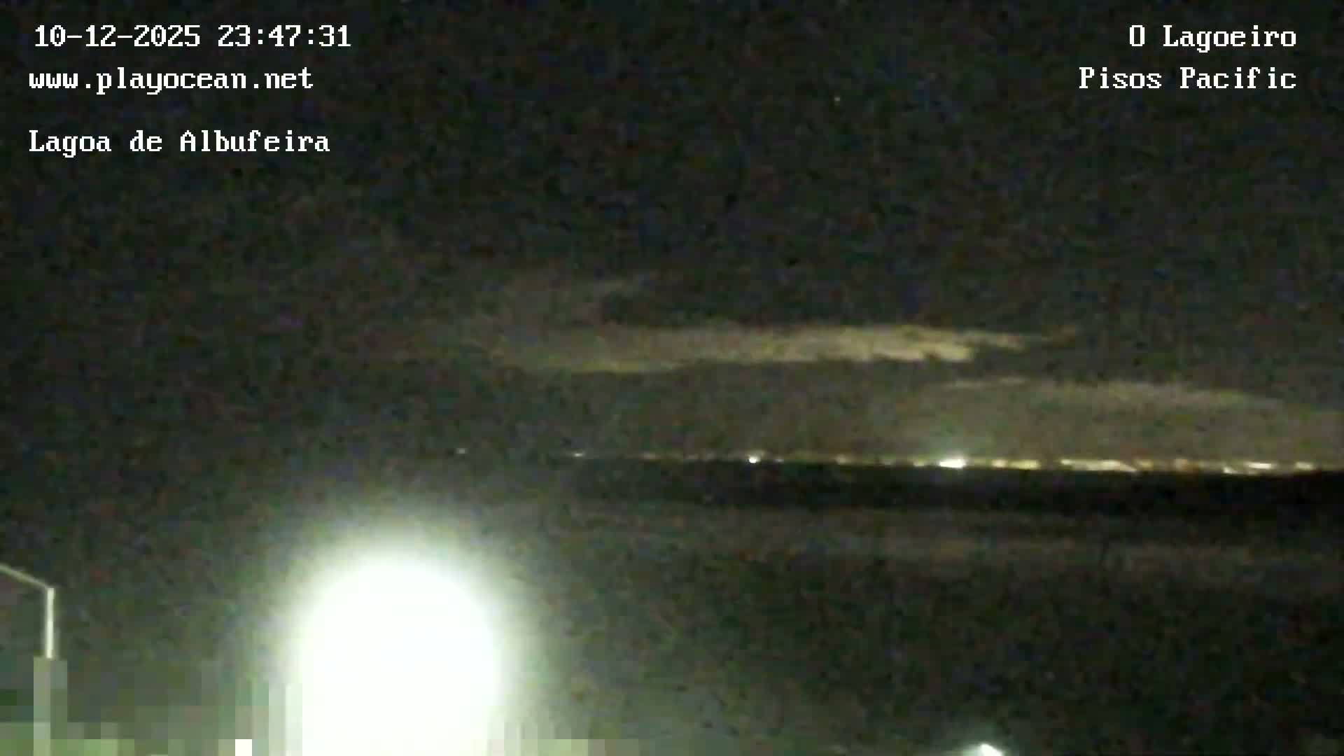 At night, a bright foreground light illuminates a blurry view of a distant city skyline across a body of water under a partly cloudy sky.