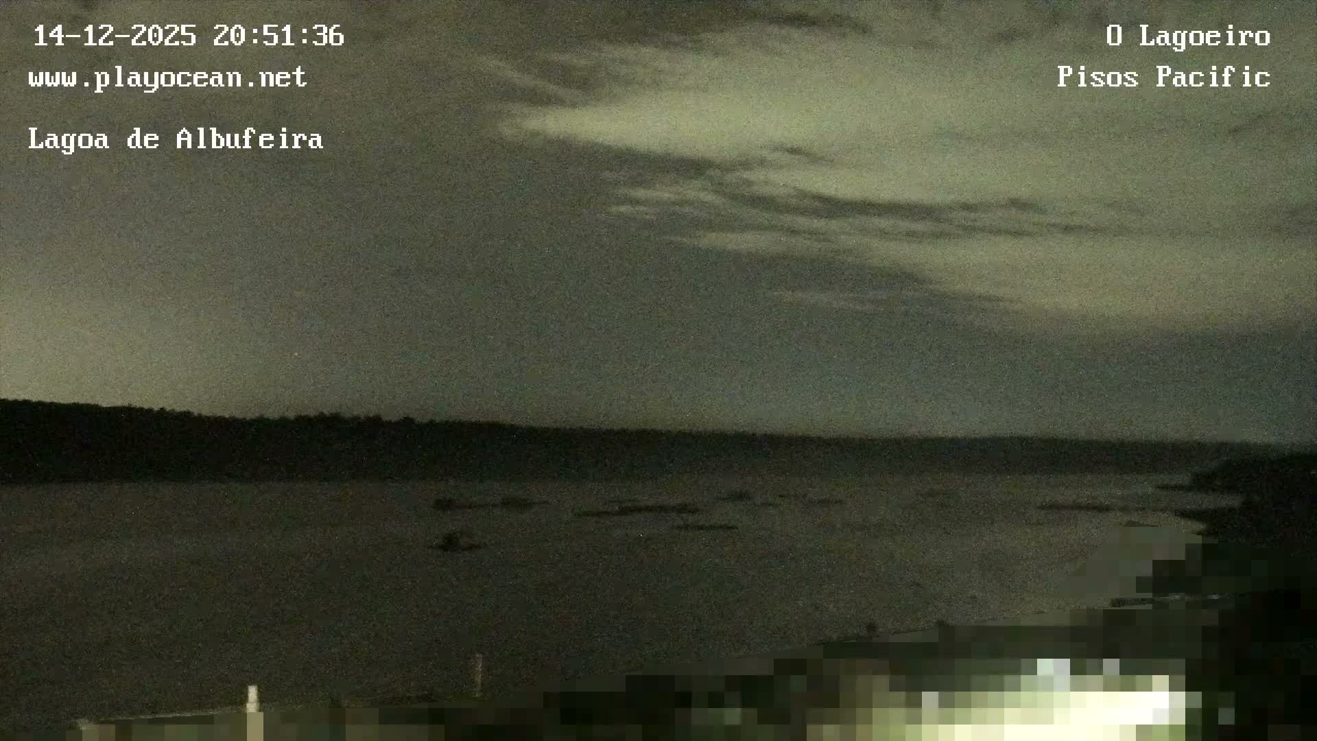 At night, a bright foreground light illuminates a blurry view of a distant city skyline across a body of water under a partly cloudy sky.