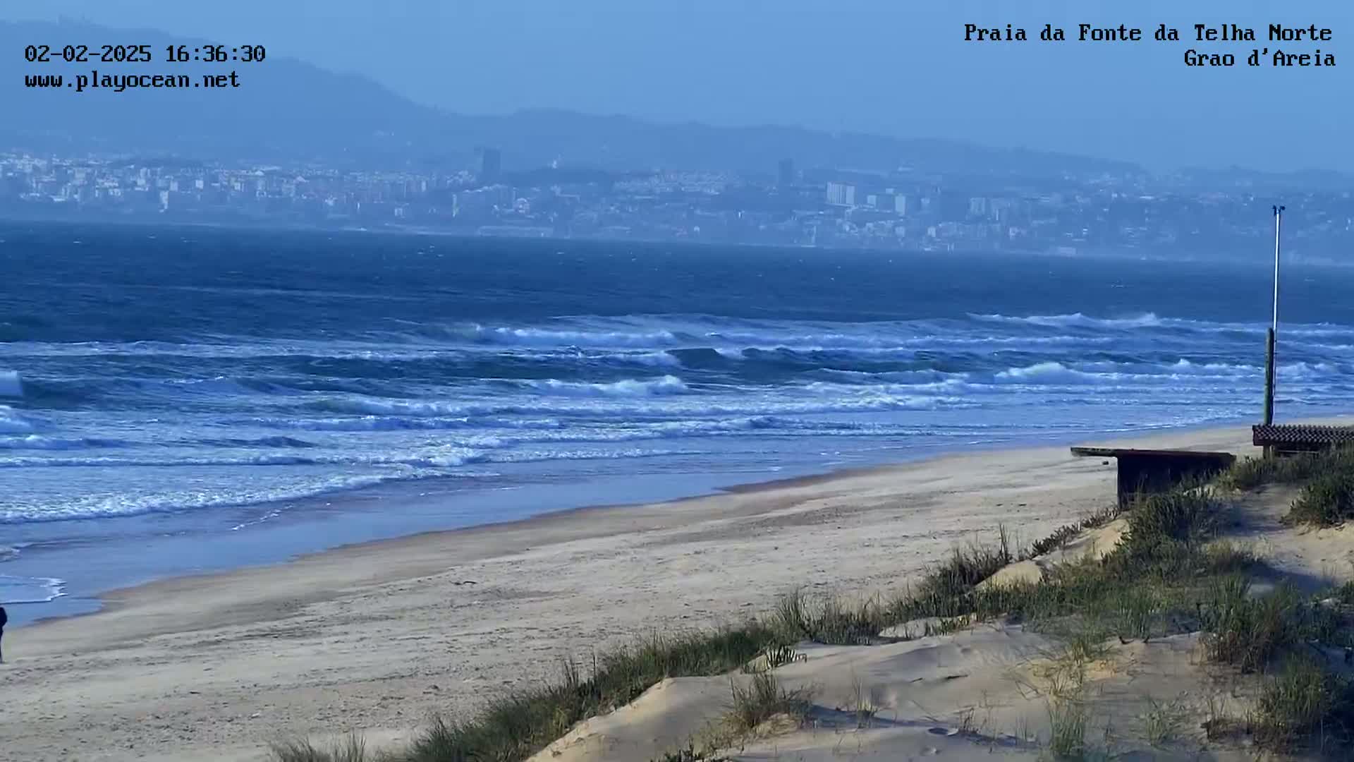 A sandy beach with moderate waves rolls onto the shore under a hazy sky, with a distant city visible in the background.