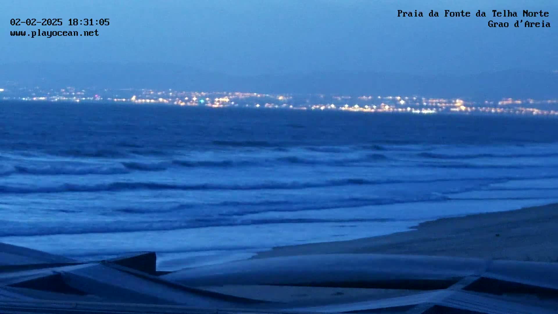 Under a twilight sky, gentle waves roll onto a sandy beach in front of a distant city skyline illuminated by lights.