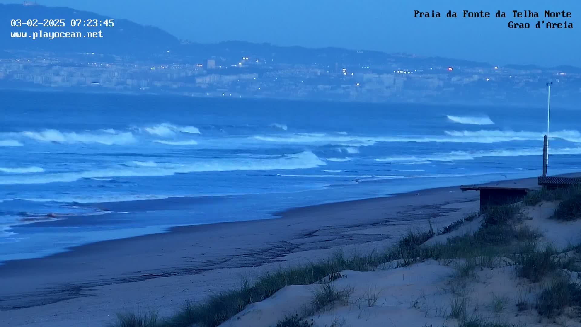 Under a twilight sky, waves roll onto a sandy beach near a distant city.