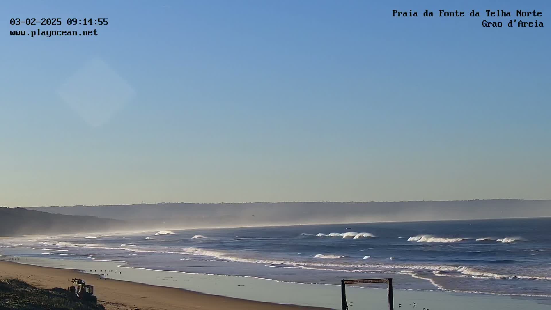 Under a clear sky, waves roll onto a sandy beach, with a hazy horizon in the distance.