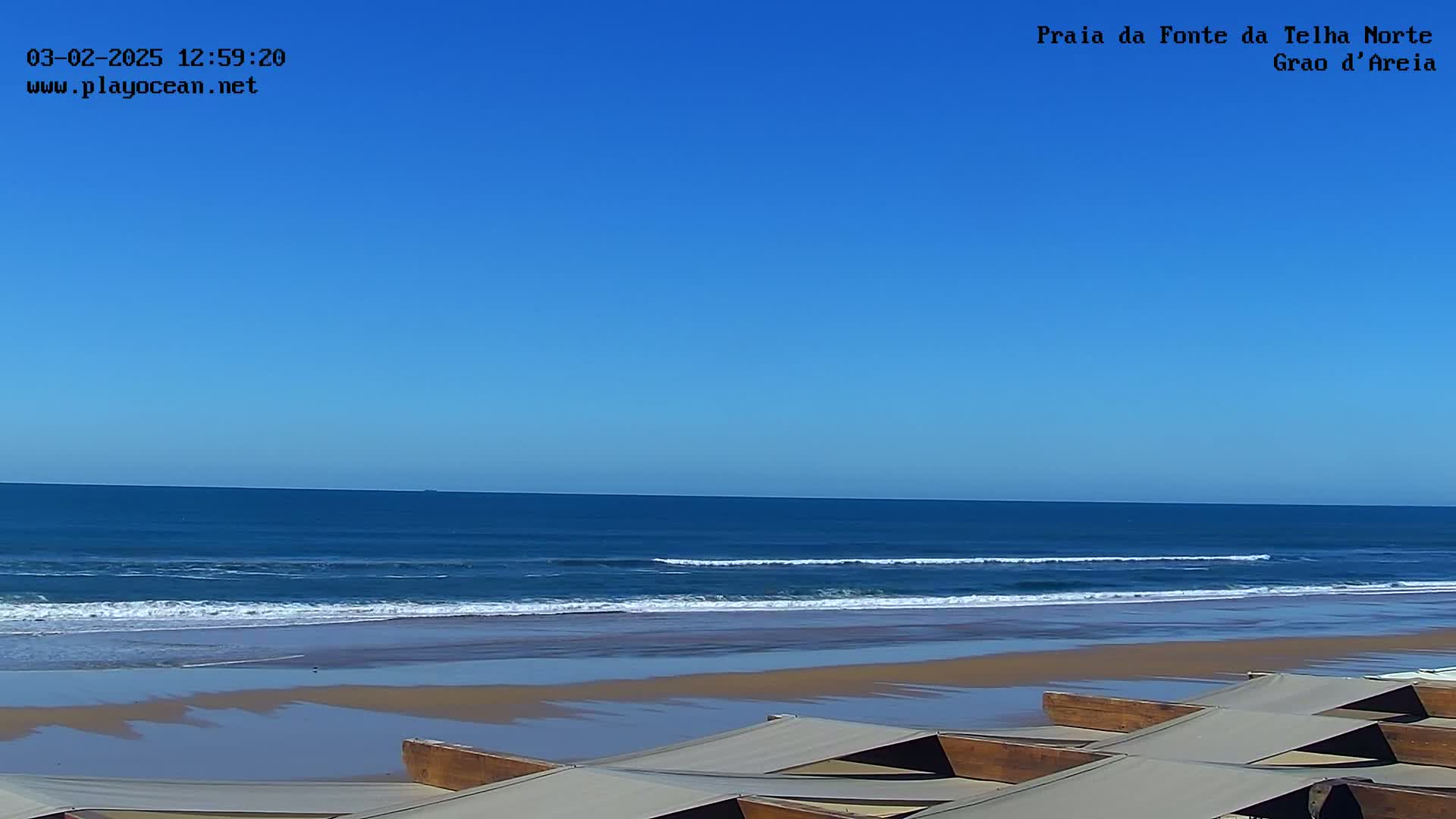 A sandy beach with calm ocean waves under a clear blue sky is partially obscured by several tan canopies.