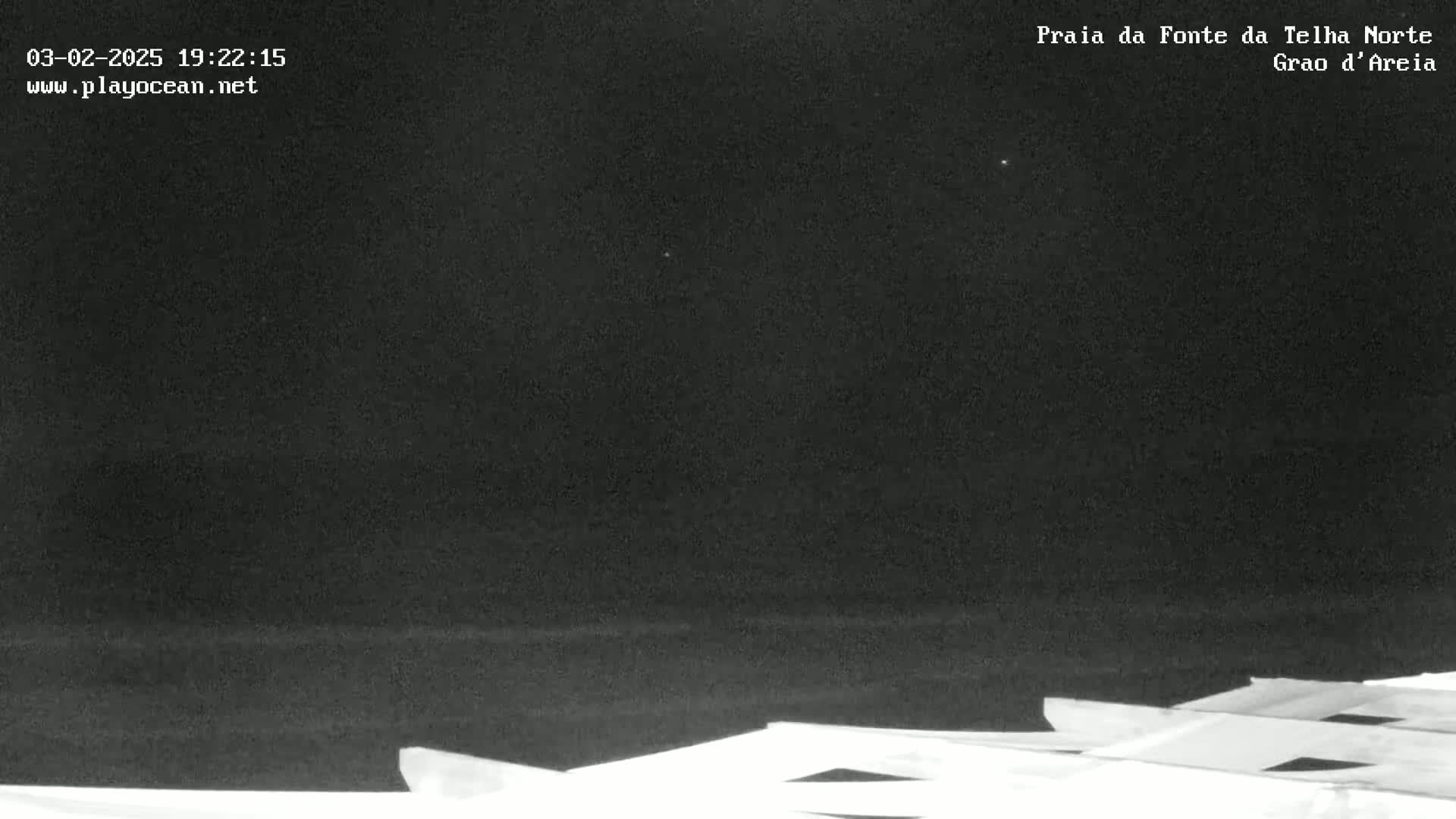 A nighttime, dark view of a beach with a few very faint stars visible in the sky, and the top of a light-colored structure at the bottom of the image.