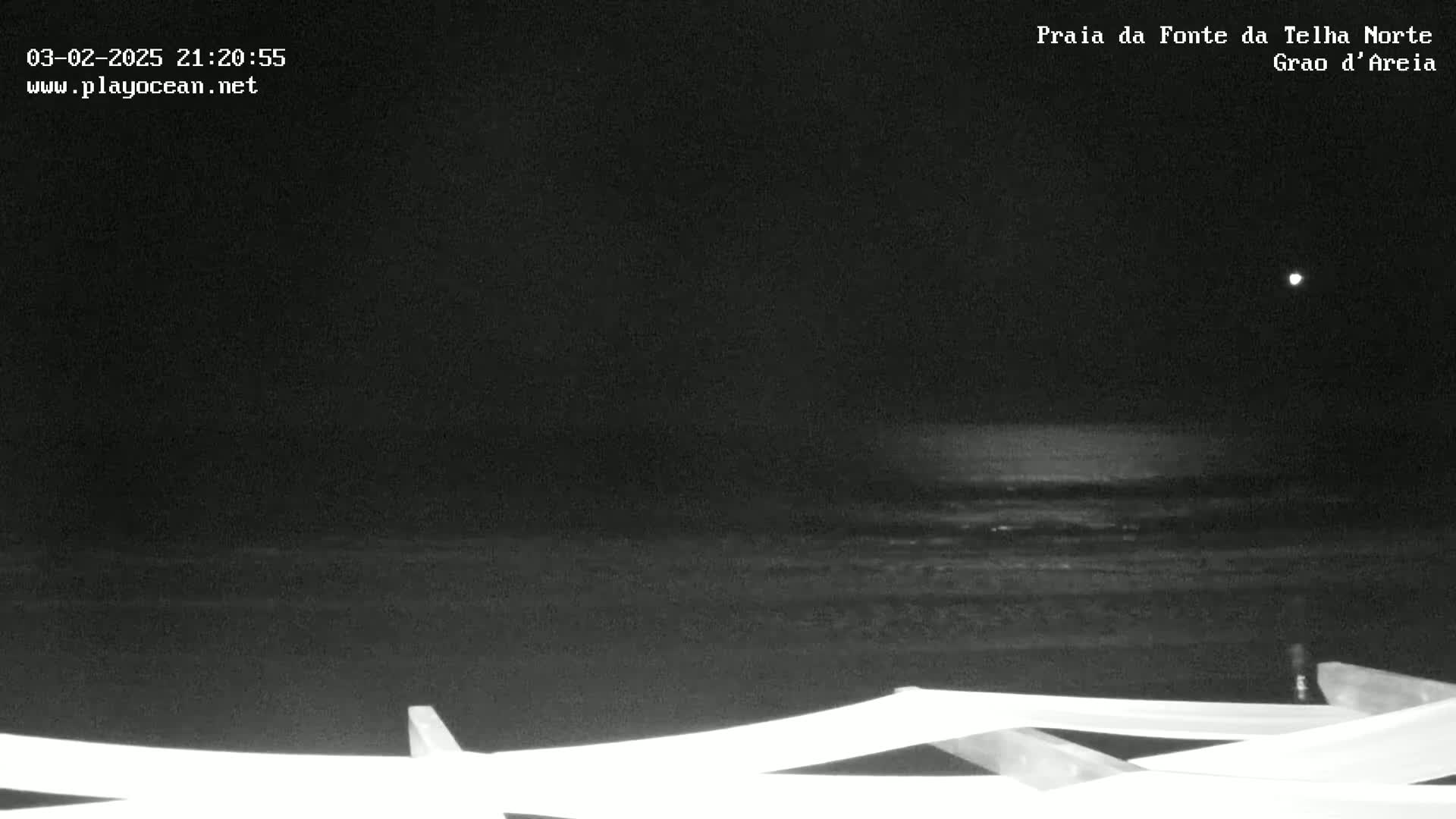 A dark nighttime beach scene shows a calm ocean under a clear sky with a bright light in the upper right corner.