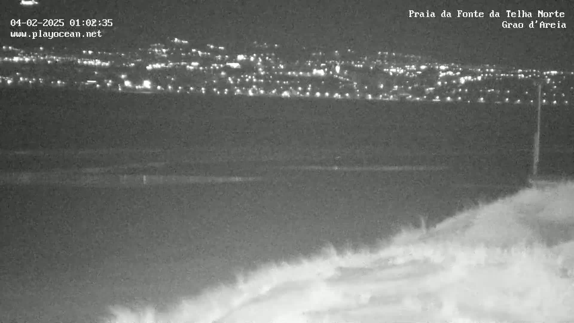 A nighttime view of a distant city's lights across a dark body of water, with a blurry foreground of what appears to be sand dunes or a beach.