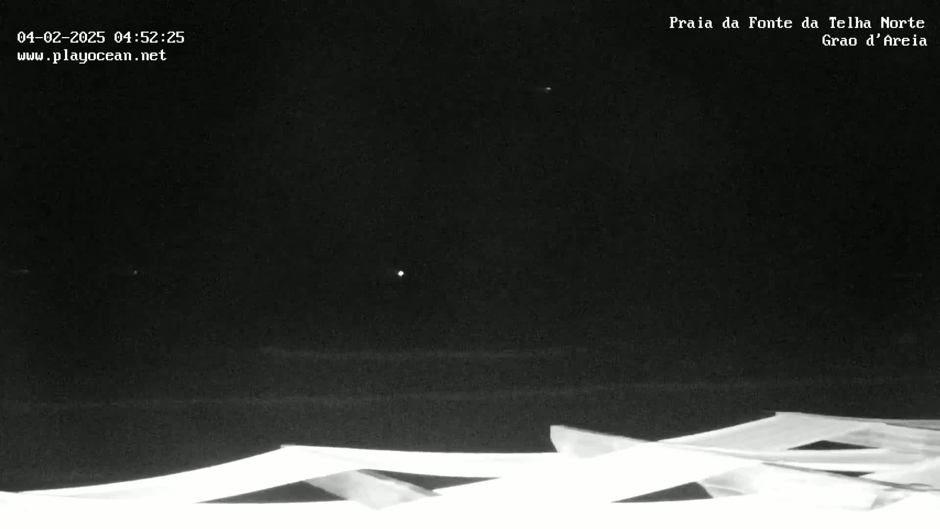 At night, a dark beach is visible beneath a mostly clear sky with a few distant lights; in the foreground are several angled, white beach chairs.