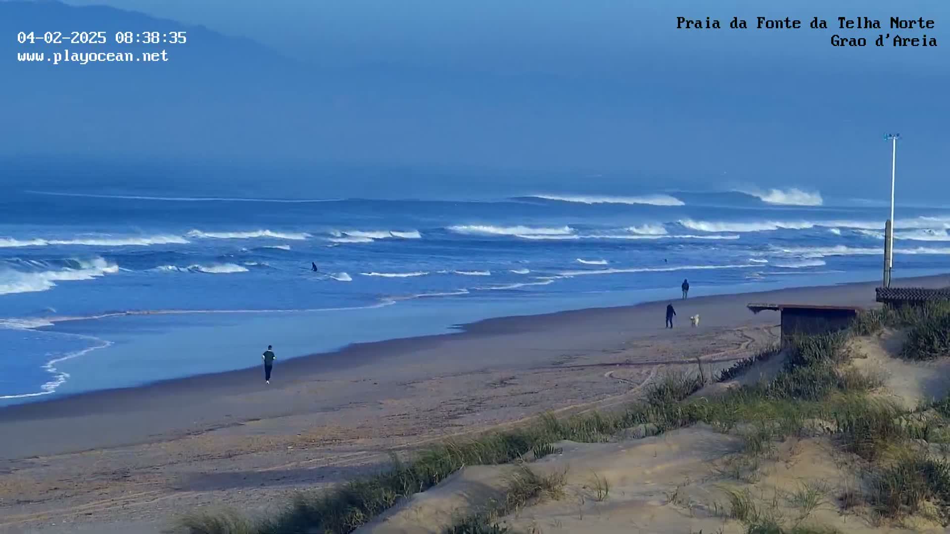 A wide sandy beach with several people walking, one person jogging, and a surfer in the ocean under a clear blue sky.