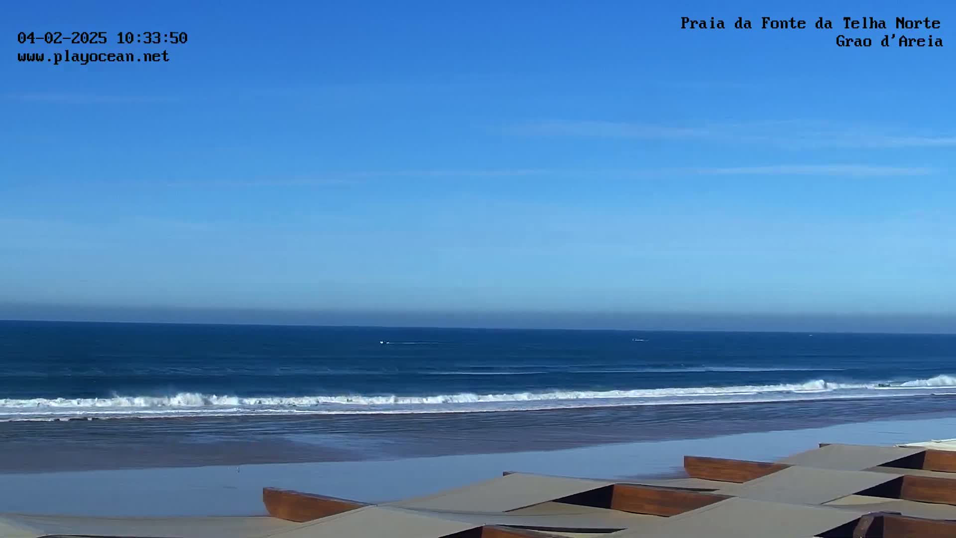 A mostly sunny day shows a wide sandy beach with gentle waves rolling onto the shore, and several wooden beach loungers in the foreground.