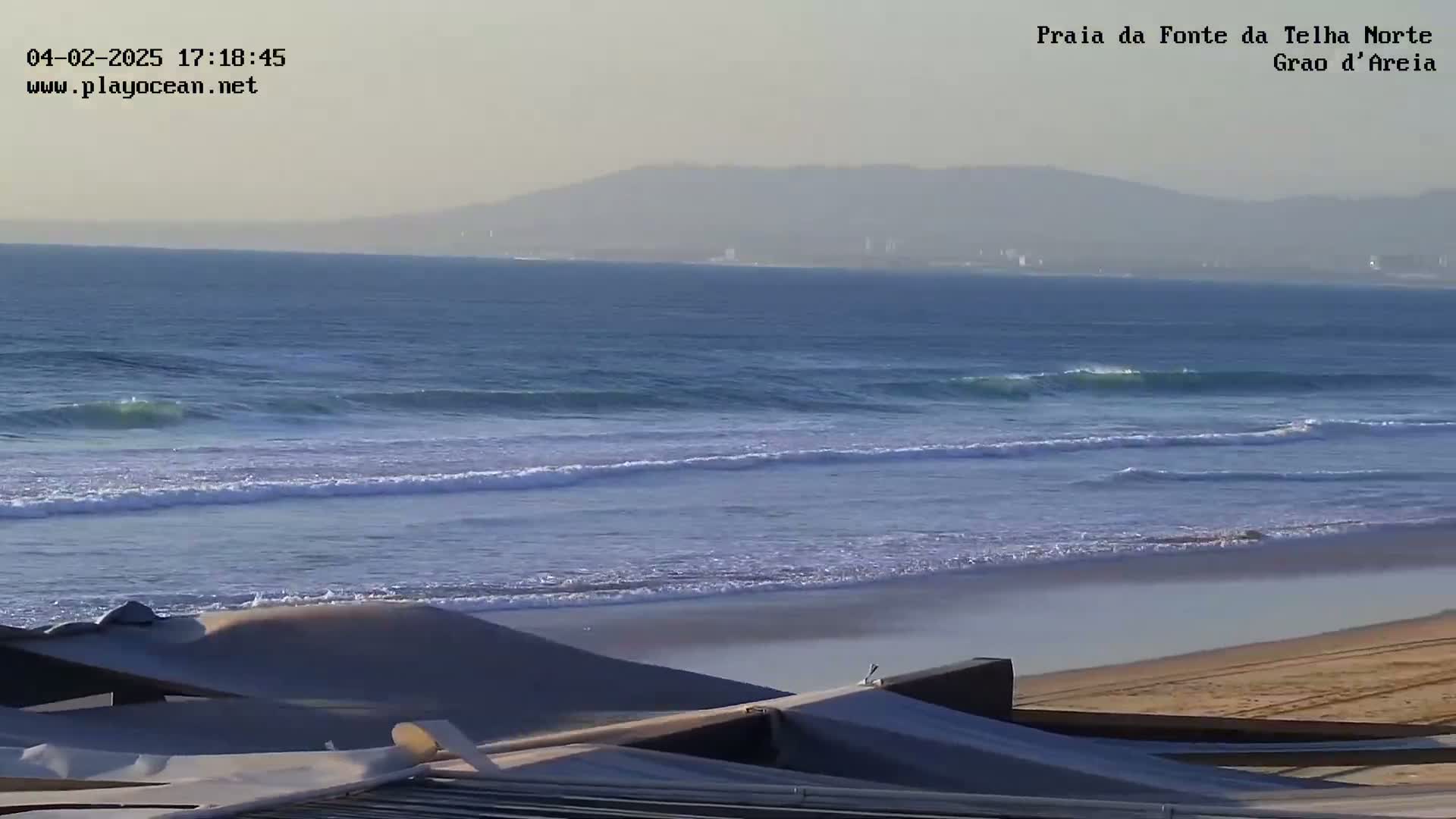 A hazy, sunny day shows gentle waves lapping a sandy beach with a distant mountain range visible on the horizon.