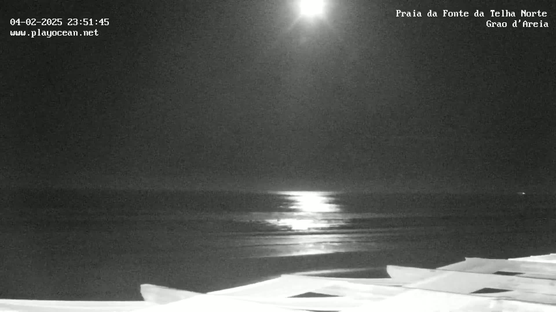 A bright moon shines over a dark, calm ocean at night, illuminating the water's surface from a beach with some structures in the foreground.
