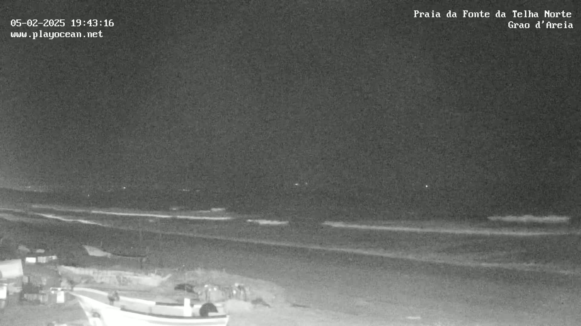 A nighttime, low-light view of a dark beach with a calm ocean and a few objects visible near the shoreline.
