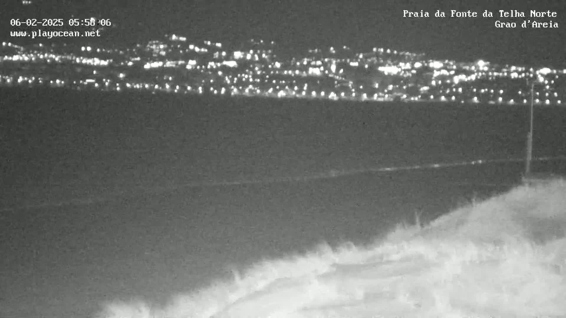 A nighttime view of a city's lights across a dark body of water, from a vantage point on a dune-like landform.