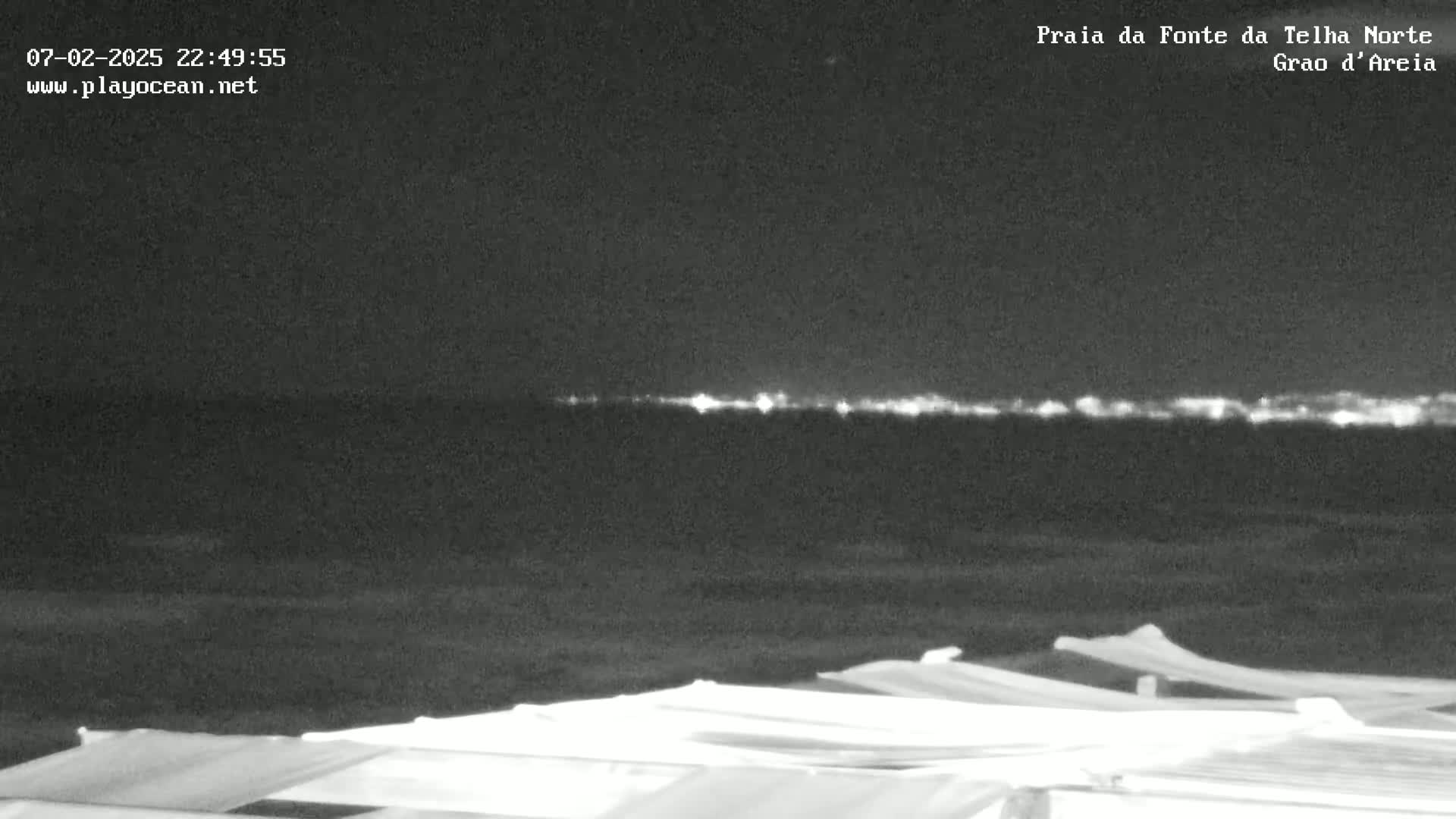 A nighttime beach scene shows a dark ocean with a faint line of lights on the horizon, and several covered structures in the foreground.