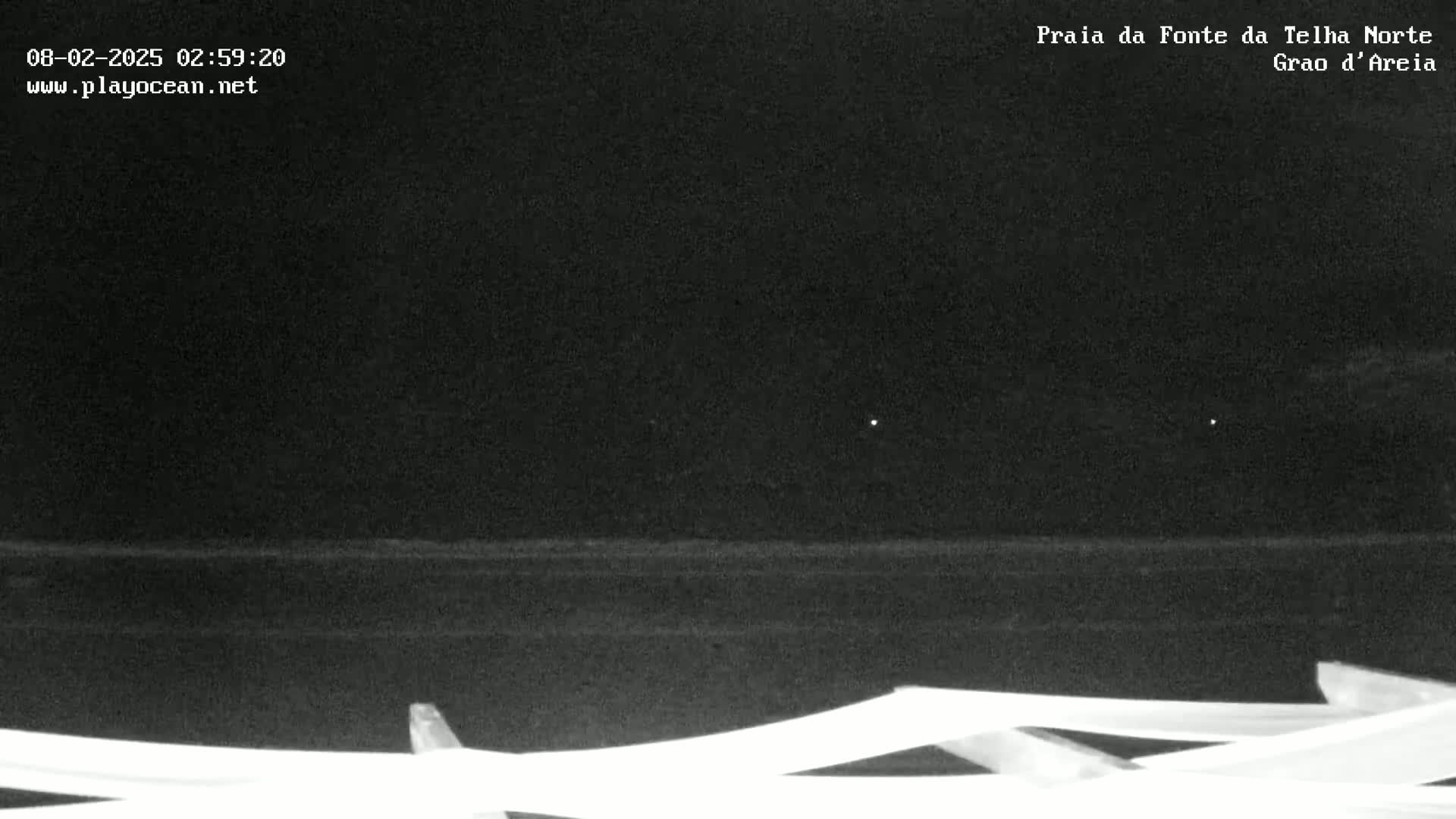 A nighttime, low-light view of a dark ocean shore with a section of white beach furniture in the foreground.