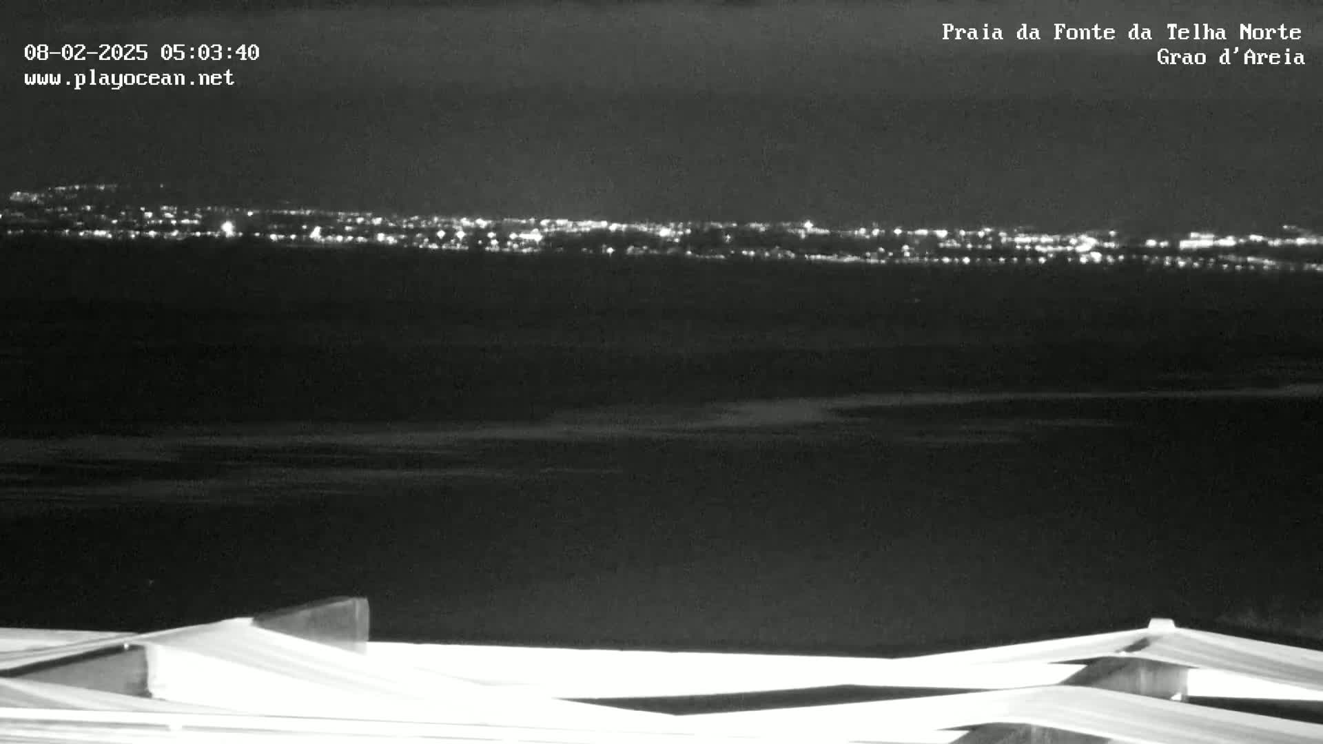 A nighttime, black-and-white view shows a distant city skyline illuminated against a dark body of water, with a structure in the foreground.