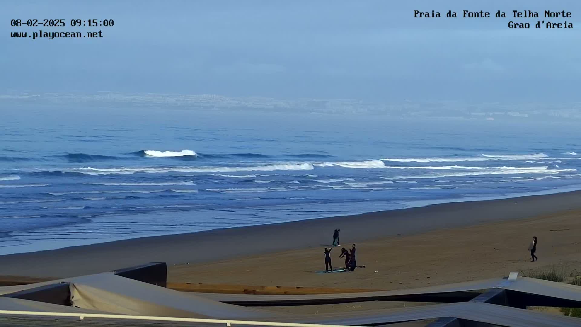 A sandy beach with several people on it is overlooked by a structure, and a hazy ocean with small waves stretches to a distant, indistinct shoreline under a clear, light-blue sky.