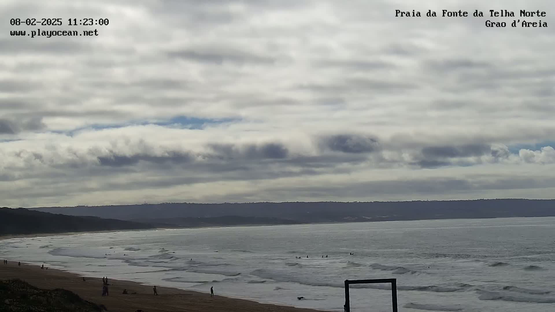 A mostly cloudy sky overlooks a sandy beach with small waves and several people, all set against a backdrop of distant hills.