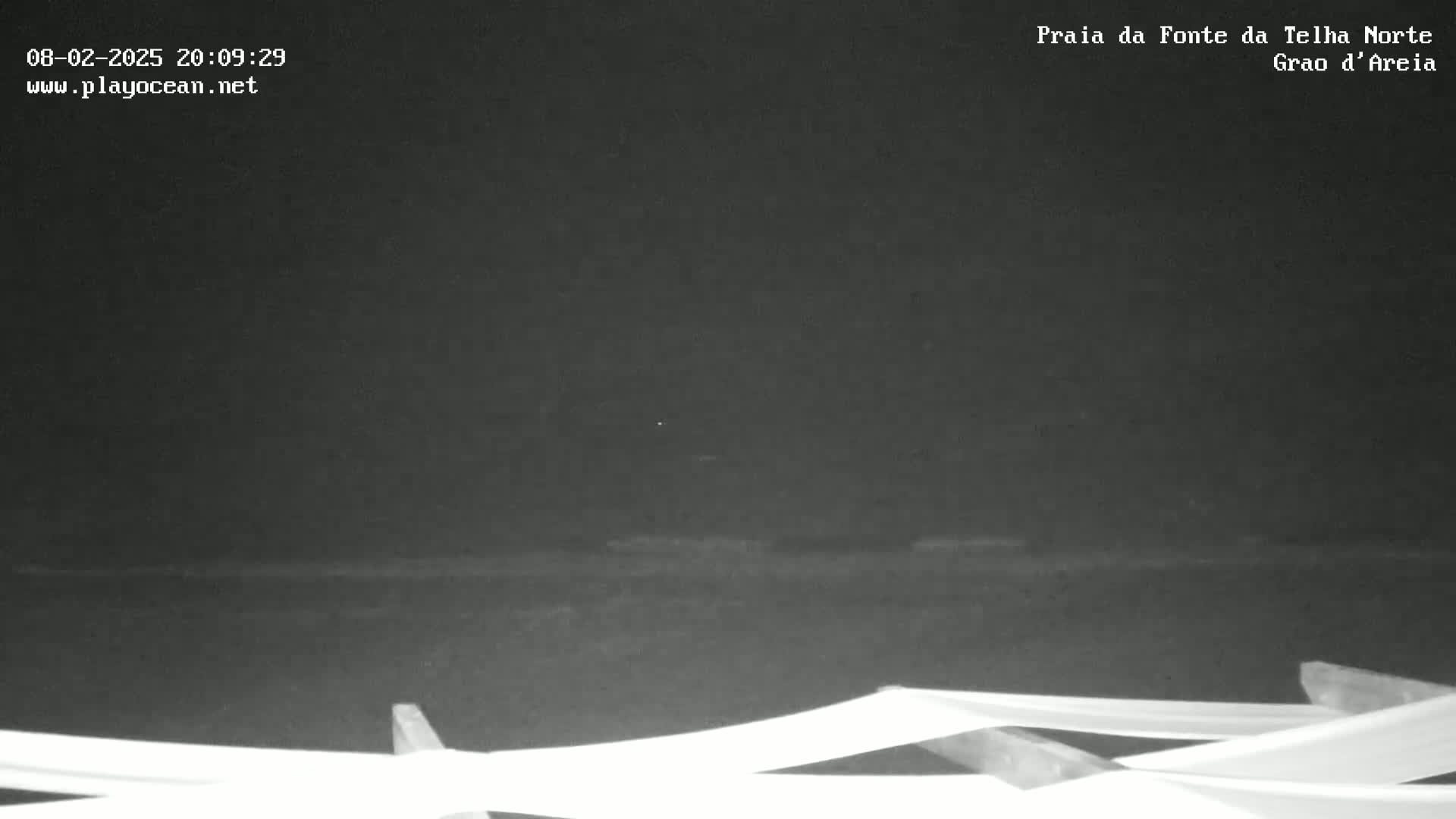 A nighttime, low-light view of a dark beach with calm water and some beach furniture in the foreground.