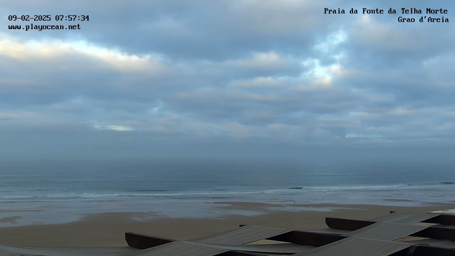 A mostly cloudy sky overlooks a calm ocean with a sandy beach and several long, low structures in the foreground.